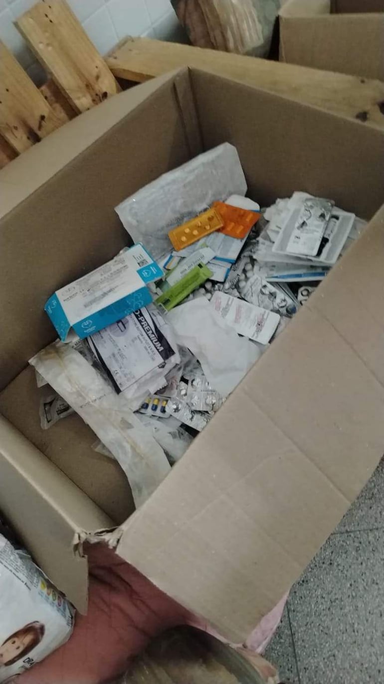 Caja de cartón llena de medicamentos y productos farmacéuticos en un ambiente sin luz natural y suelo de cerámica.