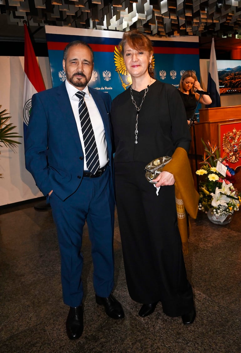 Hombre de traje azul y corbata a rayas sonríe junto a mujer con blusa negra, en un ambiente formal con decoraciones de Rusia y Paraguay.