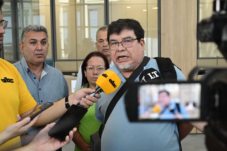 Gabriel Espínola con camiseta azul y mochila habla a micrófono. Silvio Piris y otros oyentes atentos en ambiente formal.