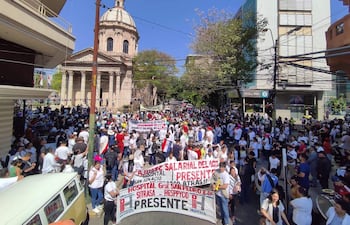 Protesta de trabajadores de la salud con camisetas blancas y pancartas, ambiente urbano y cielo despejado.