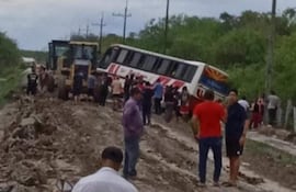 Grupo de personas, incluidos hombres con camisetas, ayudan a levantar un autobús volcado en un paisaje rural con cielo nublado.