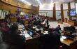 Personas sentadas en un recinto legislativo, concentradas en dispositivos móviles y documentos, en un ambiente formal con asientos rojos.
