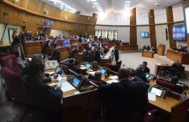 Personas sentadas en un recinto legislativo, concentradas en dispositivos móviles y documentos, en un ambiente formal con asientos rojos.