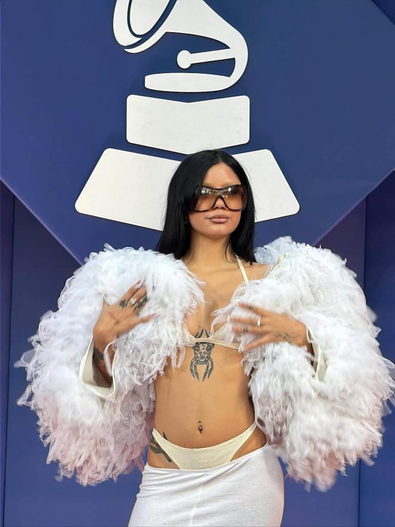 Mujer en chaqueta blanca de plumas muestra su atuendo en evento musical, con varios tatuajes visibles y fondo de logo de premio.