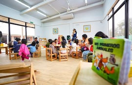 Grupo de niños con ropa colorida escucha a un adulto en aula iluminada, con un libro sobre animales en primer plano.
