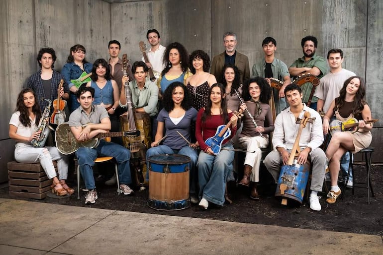 Grupo de 20 jóvenes en estudio, algunos sentados y otros de pie, sosteniendo guitarras y violines con expresiones felices.