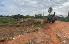 Máquinas de construcción amarillas operan en terreno rojo, con operador visible en una excavadora y cielo nublado.