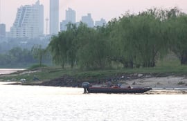 Dos personas abordo de una embarcación motorizada en el río Paraguay, con edificios al fondo y vegetación en la orilla.