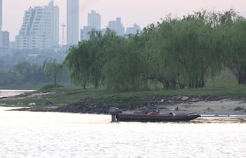 Dos personas abordo de una embarcación motorizada en el río Paraguay, con edificios al fondo y vegetación en la orilla.