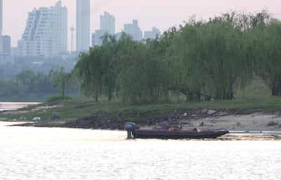 Dos personas abordo de una embarcación motorizada en el río Paraguay, con edificios al fondo y vegetación en la orilla.