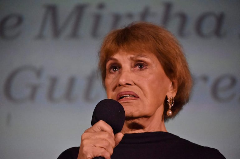 Mirtha Ibarra con camiseta negra y pendientes de perlas, sostiene un micrófono en un auditorio mientras habla con seriedad.