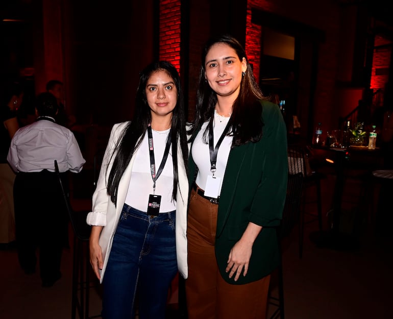 Dos mujeres con chaquetas, una blanca y otra verde, posan sonrientes en un bar con ambiente social y tenue iluminación.