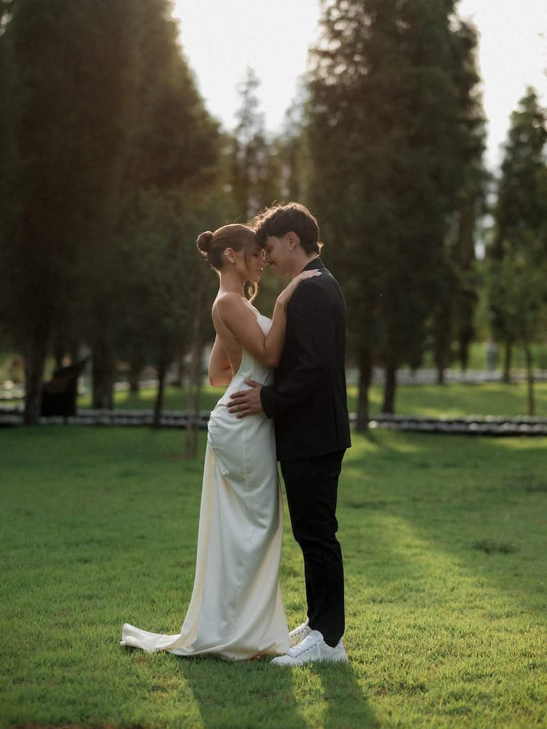 Mujer con vestido blanco y hombre en traje negro posan juntos con sonrisas, tocándose las frentes en un entorno verde.
