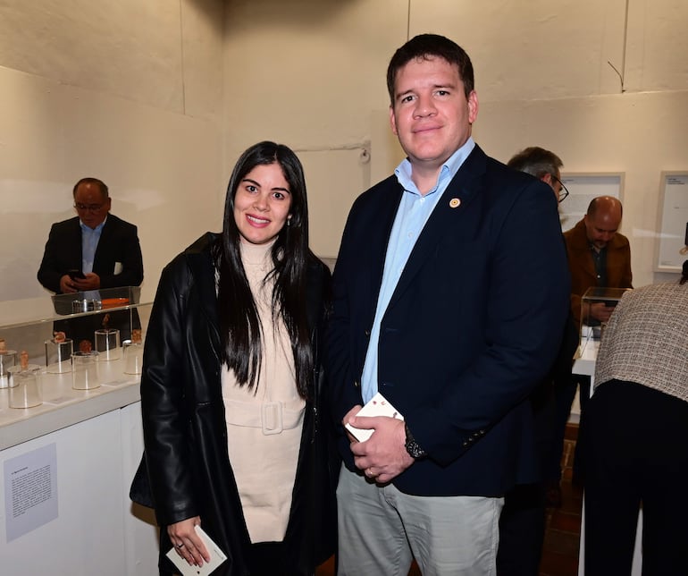 Mujer sonriente con abrigo negro junto a hombre con saco oscuro, en ambiente de inauguración de exposición cultural.
