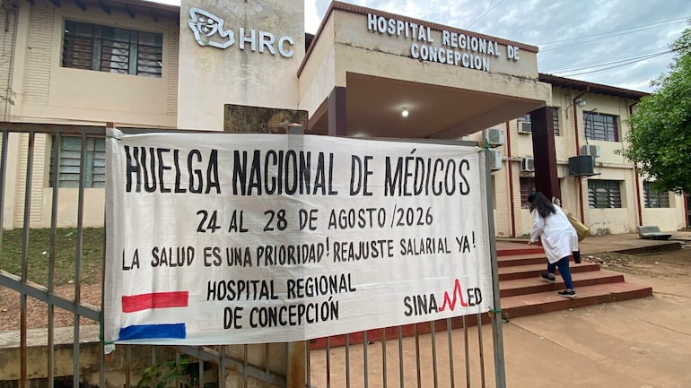 Persona en bata blanca entra al hospital con bolsa, mientras un cartel reclama por la salud y salarios.