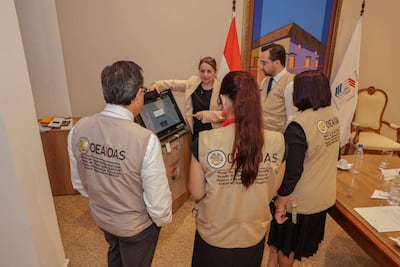Mujer de cabello largo explica mientras apunta a una máquina, rodeada de observadores con chalecos de la OEA en sala de reuniones.