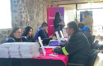 Cuatro mujeres en camisetas azules interactúan con un hombre de abrigo oscuro en una feria de empleo, rodeados de mesas con documentos.