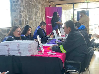 Cuatro mujeres en camisetas azules interactúan con un hombre de abrigo oscuro en una feria de empleo, rodeados de mesas con documentos.