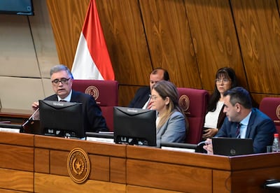 Miguel Baez Reyes con gafas, vestido formal, habla en mesa con otros presentes atentos y documentos alrededor, en un ambiente institucional.