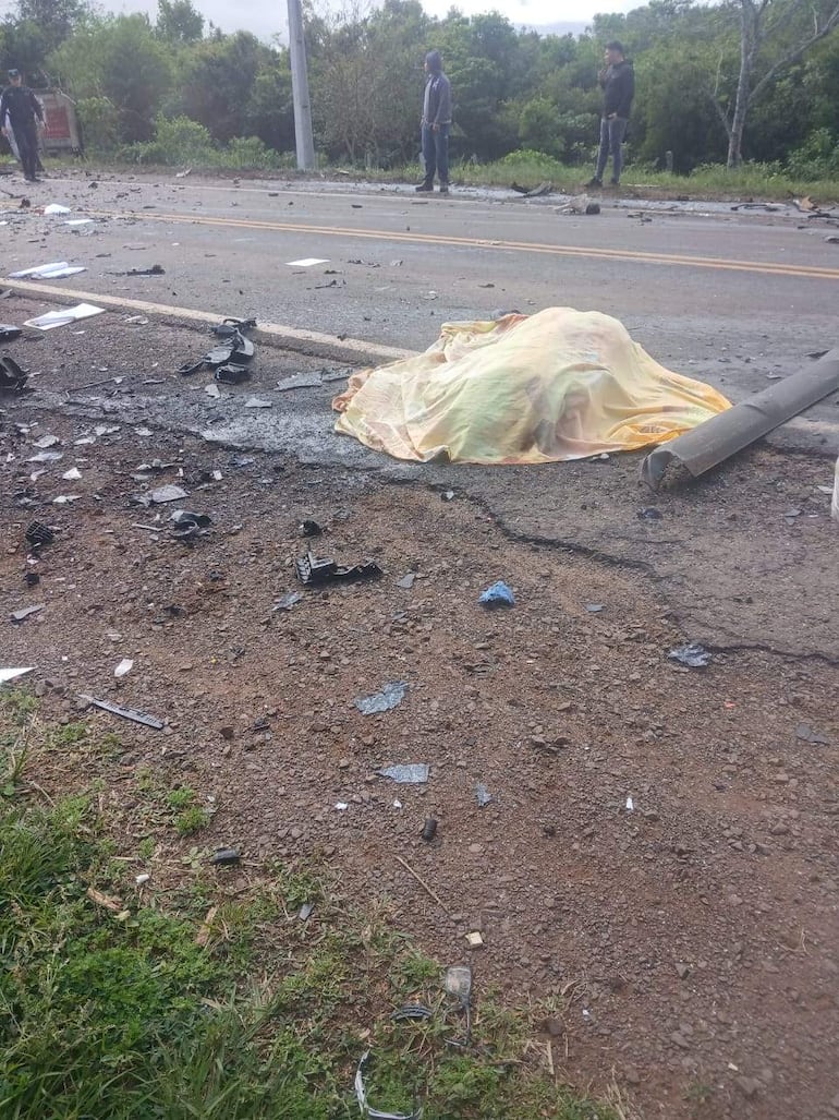 Tres personas de pie, una en posición de observación, junto a un objeto cubierto por una manta en una carretera rodeada de vegetación.
