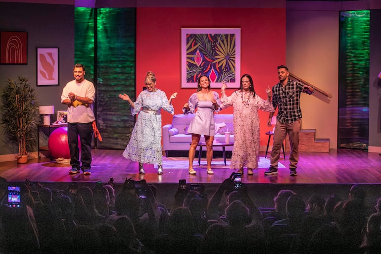 Cuatro mujeres y un hombre en el escenario, vestidos coloridamente, expresando alegría mientras el público los fotografía.