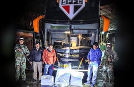 Cinco personas, dos en uniformes militares y uno en abrigo naranja, junto a un autobús 'Cosmos' con paquetes en el suelo.