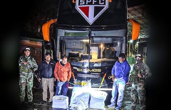 Cinco personas, dos en uniformes militares y uno en abrigo naranja, junto a un autobús 'Cosmos' con paquetes en el suelo.