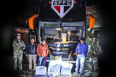 Cinco personas, dos en uniformes militares y uno en abrigo naranja, junto a un autobús 'Cosmos' con paquetes en el suelo.