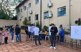Niño con camiseta a rayas, adultos conversando, mujer en mesa informativa. Ambiente de actividad electoral al aire libre.