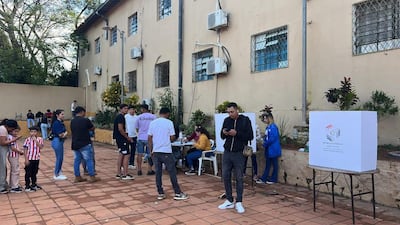 Niño con camiseta a rayas, adultos conversando, mujer en mesa informativa. Ambiente de actividad electoral al aire libre.