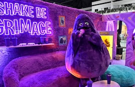Figura de Grimace, color púrpura, sonriente, sostiene un vaso en un ambiente colorido con letrero luminoso y vasos sobre mesa blanca.