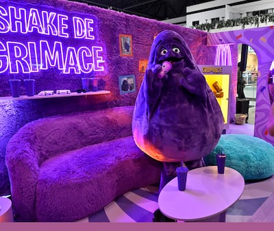 Figura de Grimace, color púrpura, sonriente, sostiene un vaso en un ambiente colorido con letrero luminoso y vasos sobre mesa blanca.