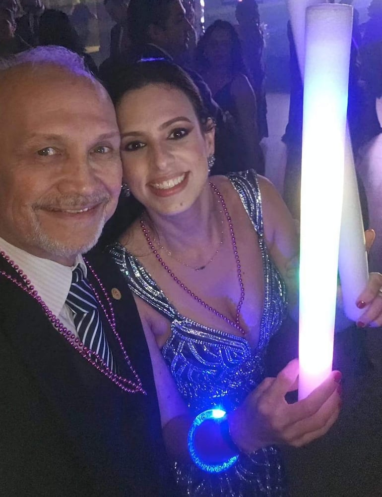 Mario Ferreiro con traje negro y cadena morada, sonríe junto a mujer de vestido brillante con pulsera iluminada en ambiente festivo.