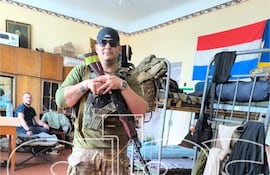 Eduardo Román vestido con uniforme militar sostiene un fusil, junto a un compañero sentado en una base con bandera paraguaya.