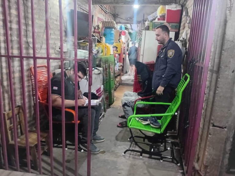 Dos hombres en sillas, uno con camiseta negra y bandera paraguaya escribe, el otro con chaqueta de seguridad escucha atentamente.