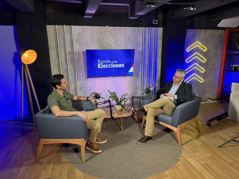 Alvaro Grau en saco negro habla con un hombre en camiseta verde, en un estudio con planta y micrófono.