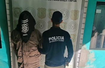 Hombre con capucha de tono marrón es detenido por agente policial con chaqueta negra en sede policial.