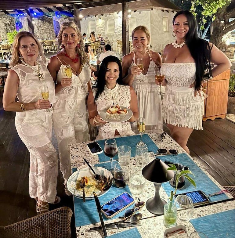 Cinco mujeres vestidas de blanco sonriendo alegremente, sosteniendo copas de bebida junto a una tarta decorada en un ambiente festivo.