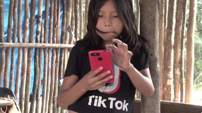 Niña con cabello largo, camiseta negra con logo de TikTok, sosteniendo un teléfono móvil rojo en un entorno de madera simple.