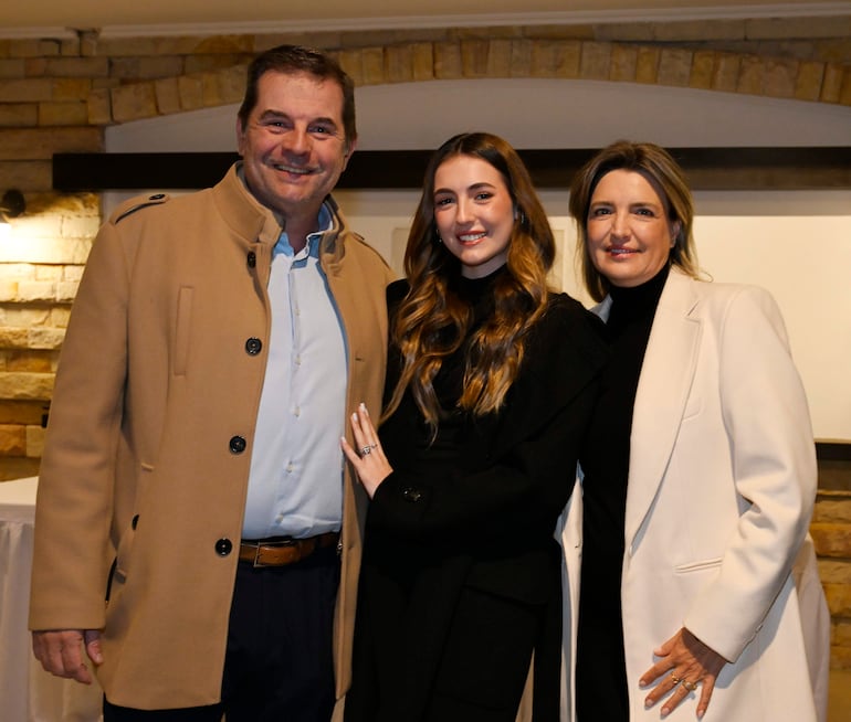 Hombre en abrigo claro y mujer en negro sonríen junto a otra mujer en abrigo blanco en ambiente formal.