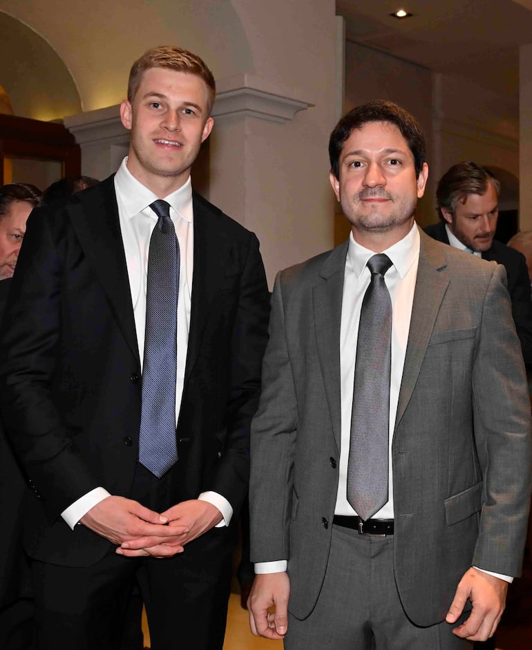 Raúl Valdez, con traje gris y corbata, junto a un hombre rubio en traje negro, durante un evento formal en el Hotel La Misión.