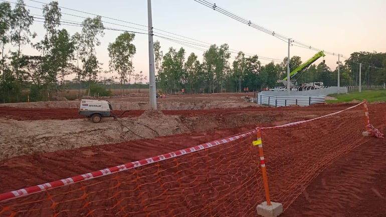Un sitio de construcción con maquinaria pesada levantando estructuras metálicas y trabajadores de naranja al fondo en terreno excavado.