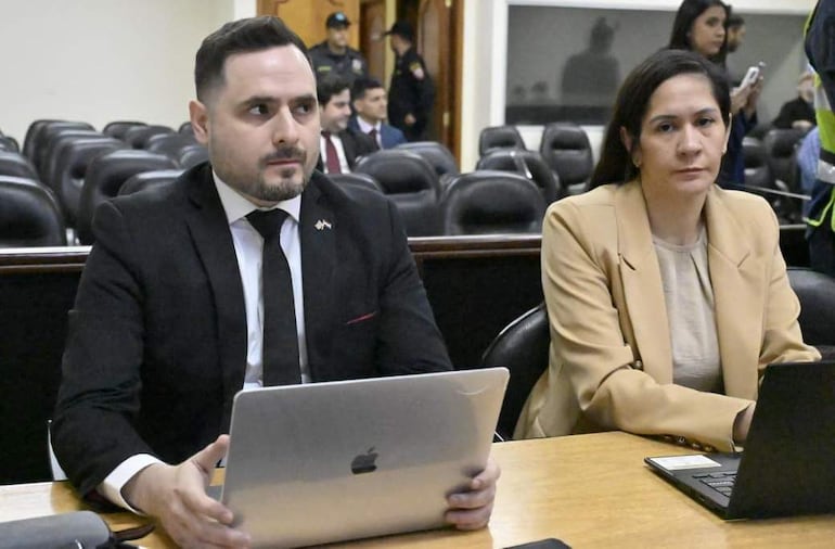 Corbeta en traje negro y Valdez en blazer beige, ambos con laptops, en ambiente formal de tribunal.