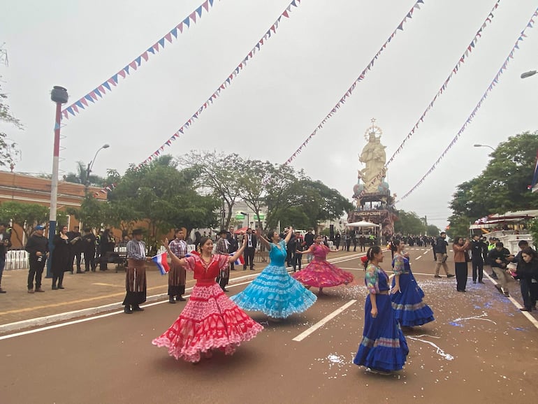 Bailarinas con trajes coloridos sostienen banderas de Paraguay, ambiente festivo con confeti y gran estatua religiosa al fondo.