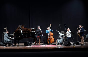 Cinco músicos en el escenario: pianista, cantante, bajista, baterista y saxofonista, iluminados con luces tenues en fondo negro.