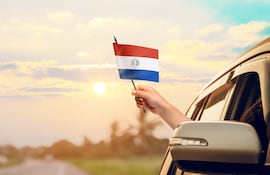De la ventanilla de un automóvil circulando por una ruta sale una mano que sostiene una bandera paraguaya.