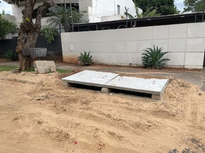 Área de construcción con foso cubierto por losas de cemento, rodeado de arena suelta y vegetación en un entorno urbano.