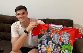 Hombre joven sentado en sofá, sosteniendo camiseta blanca mientras sonríe, en un ambiente decorado con fotos futbolísticas.