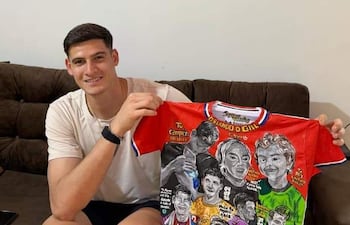 Hombre joven sentado en sofá, sosteniendo camiseta blanca mientras sonríe, en un ambiente decorado con fotos futbolísticas.
