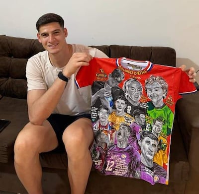 Hombre joven sentado en sofá, sosteniendo camiseta blanca mientras sonríe, en un ambiente decorado con fotos futbolísticas.